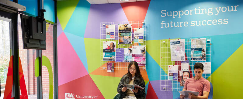 Two students sit in a hallway in front of a brightly decorated wall. There are posters and flyers on the wall behind them and the slogan 'supporting your future success.